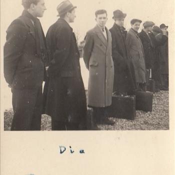 Foto van 8 mannen op een rij, in burgerkleding met een koffer