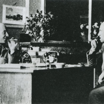 Foto van twee mannen zittend aan weerszijden van ee bureau met twee telefoons. Beide mannen roken een sigaar.