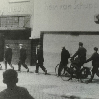 Twee Duitse militairen (een op fiets) brengen vier Nederlandse burgers op. Op achtergrond winkels Albert Heijn en Hein van Schuppen. Tekst achterop: "Terreur".