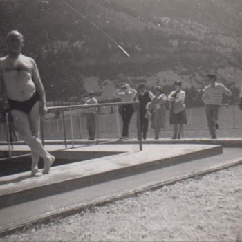 Foto van een man in zwembroek leunend tegen een omheining bij een zwembad.